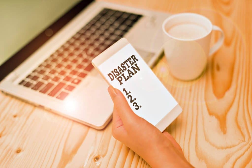 Person reviewing disaster plan on smartphone beside laptop