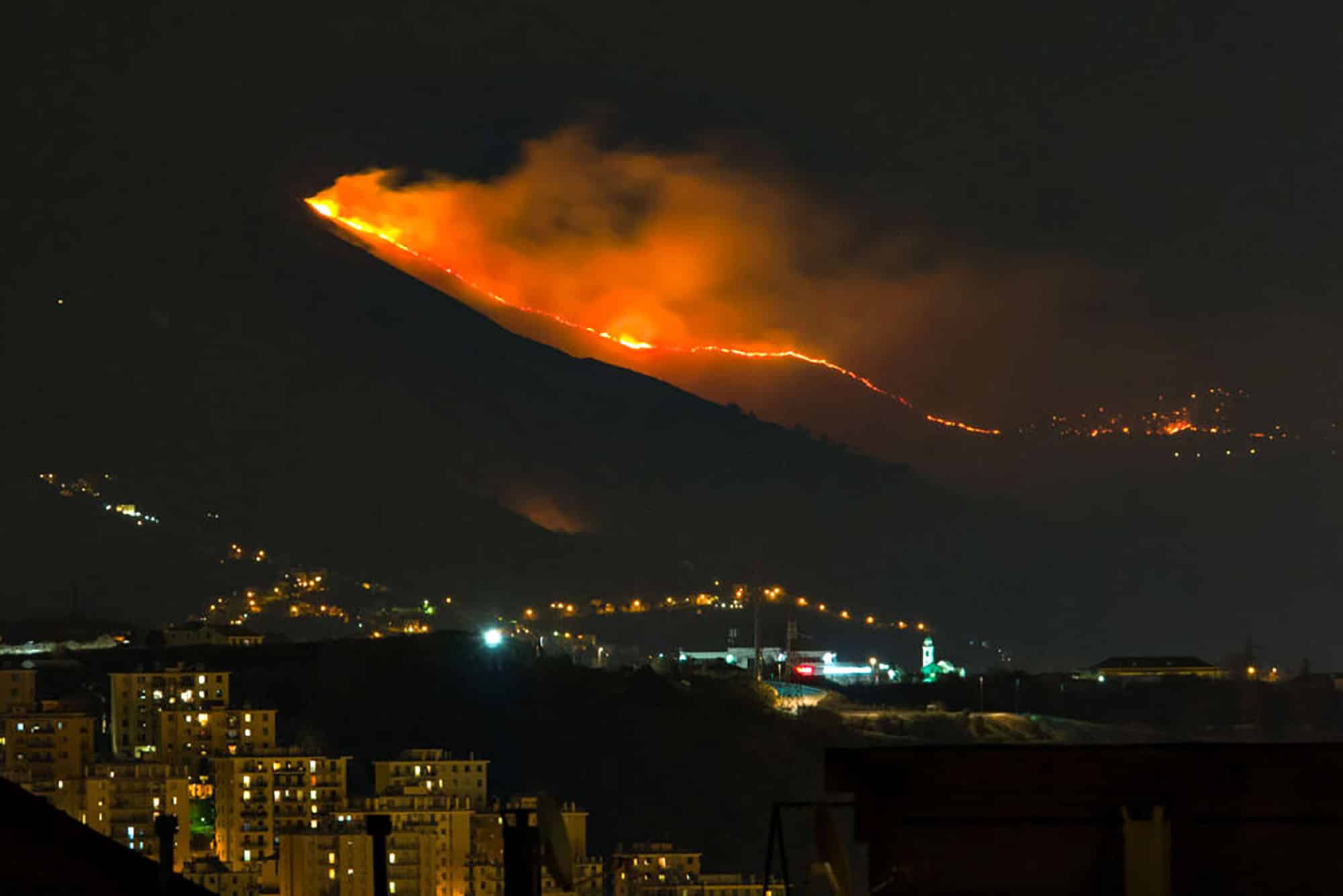 Nighttime hillside wildfire near city, smoke illuminated by flames