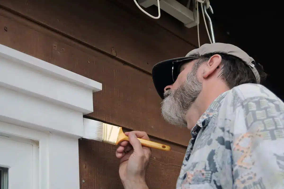 Man painting wooden house exterior near window trim with brush